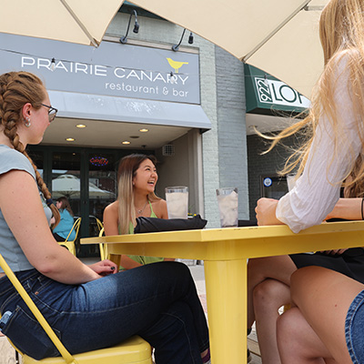 Three girls sitting at a table outside of a restaurant called "Prairie Canary". The girls are laughing.