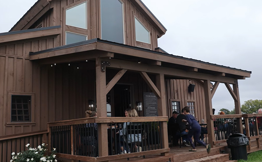 A barn with a patio and people sitting and talking. A child is running up the stairs.