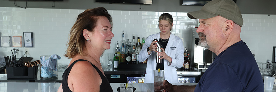 Two people talking at a bar while the bartender makes a drink