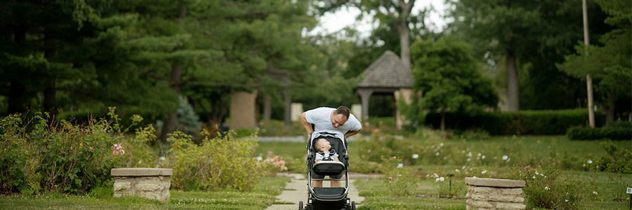South of Grand a father pushing a stroller with a baby down a path surrounded by greenery. The baby and father are both smiling.