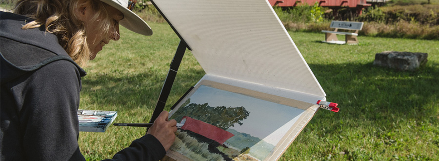 Paint Madison County A woman in painting a covered bridge with the covered bridge in front of her.