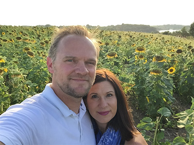 A couple smiling at the camera in front of a field of sunflowers.