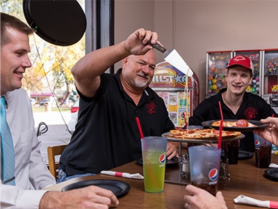 Three men sitting at a table looking at pizza. One of the men is getting a slice.