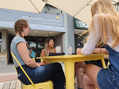Prairie Canary Three young women sitting at a table outside of a cafe laughing.