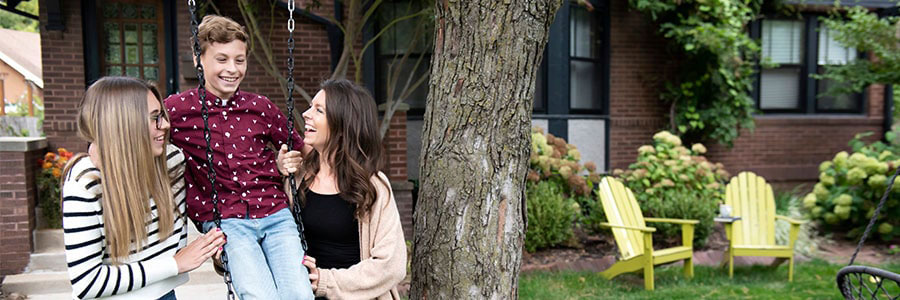 two women holding a boy on a swing in front of a brick house